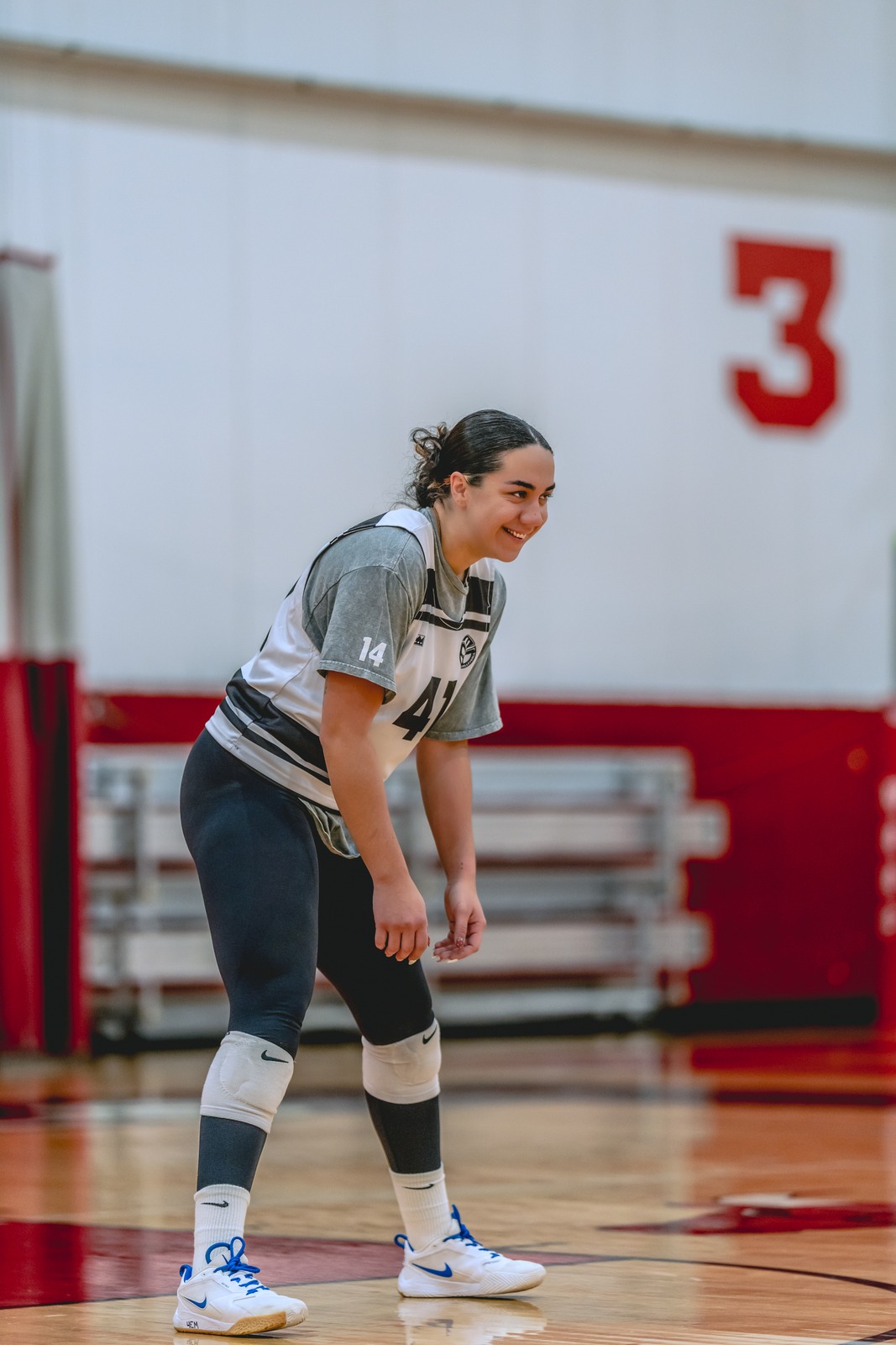 A player in a gray top and dark leggings crouches on a wooden court.