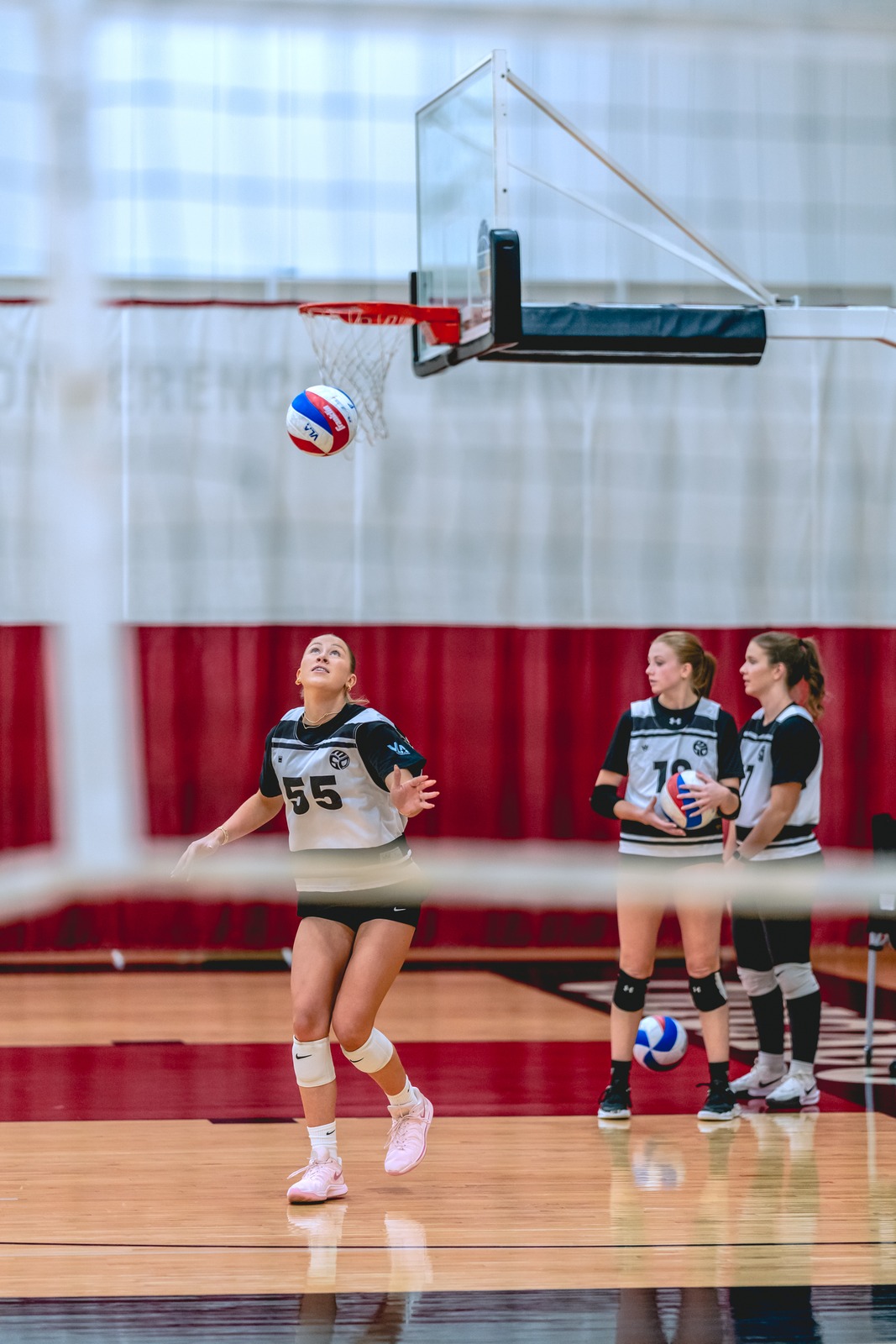 A player in a white and black top looks up as a volleyball flies overhead. Two other players in white and black tops stand nearby.
