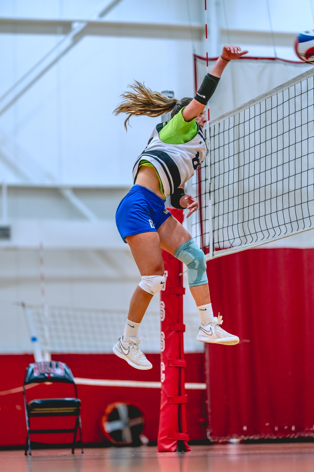 A player in a white and green top and blue shorts jumps with her arm extended.