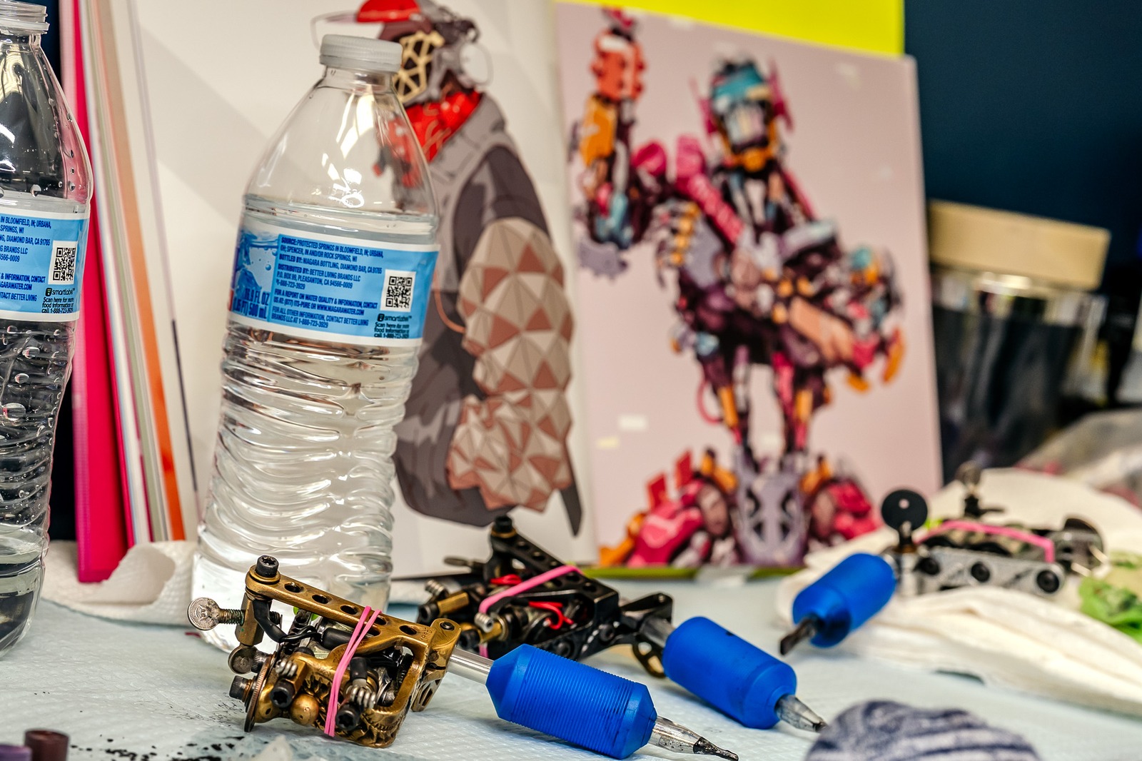 Tattoo machines and water bottles are on a table with artwork in the background.