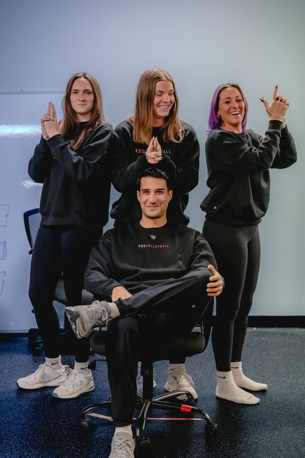 Four people in black tops pose around a chair.