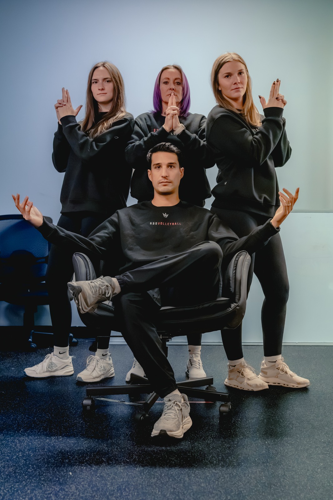 Four people in black tops pose with hand gestures around a chair.