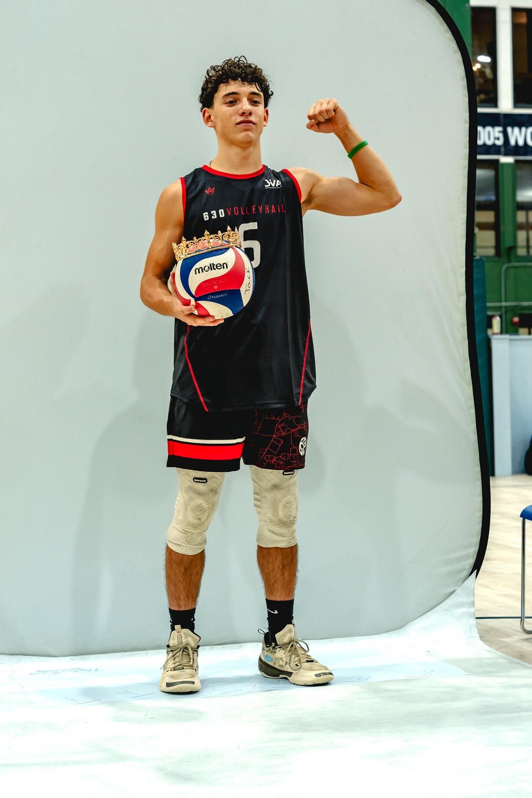 A player in a black top and shorts holds a volleyball and flexes his arm.