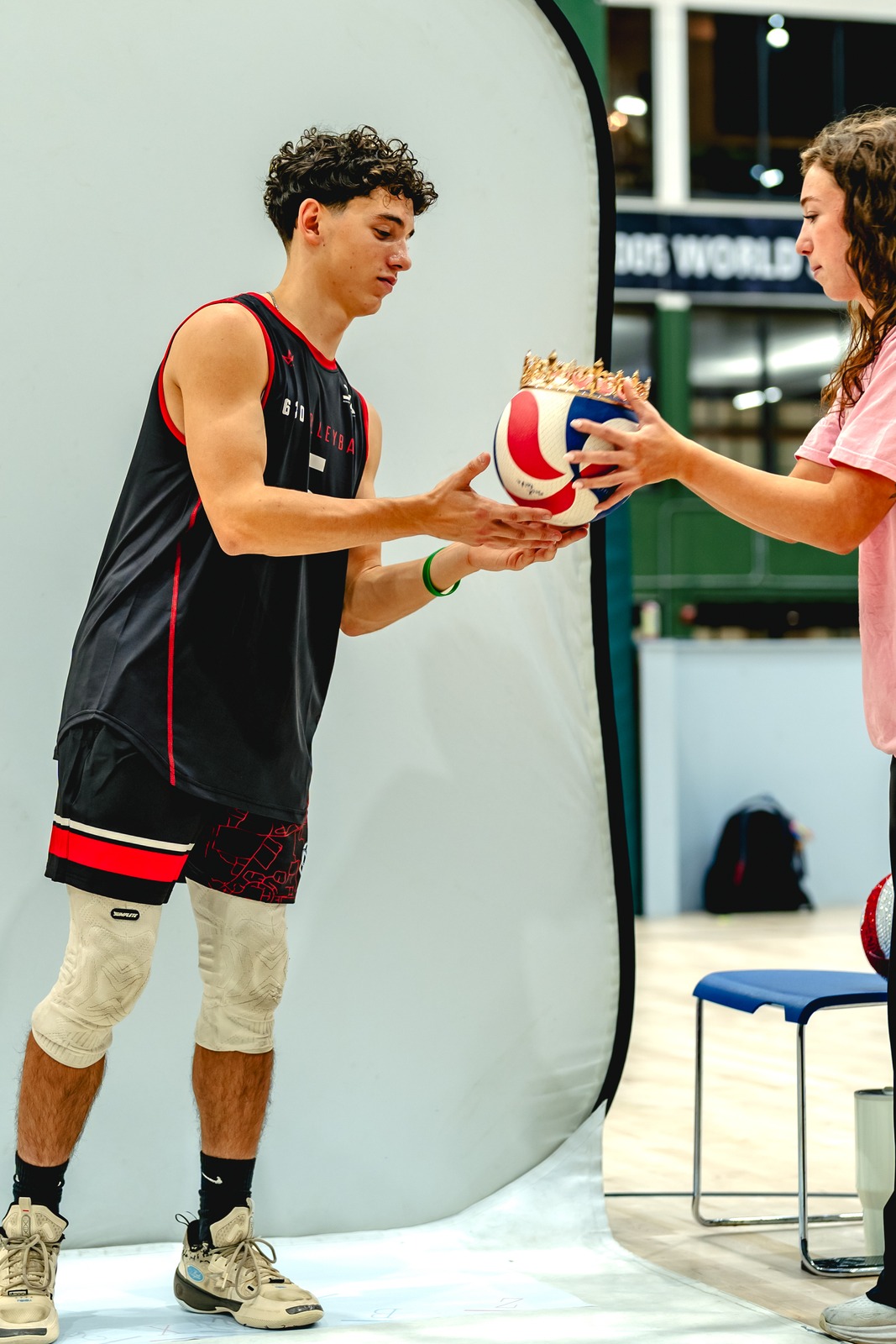 A player in black and a person in pink hold a crowned volleyball beside a photo backdrop.