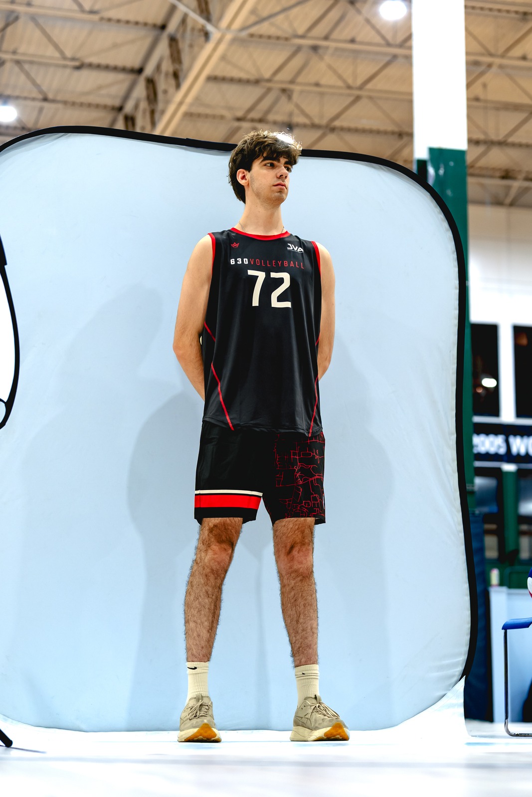 A player in a black and red uniform stands facing forward with hands clasped behind their back, looking slightly to the right.