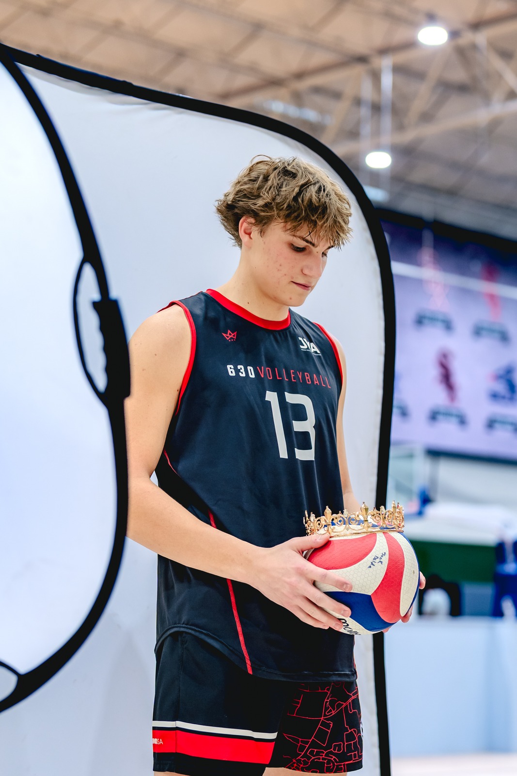 A player in a black top and shorts holds a volleyball and a crown.
