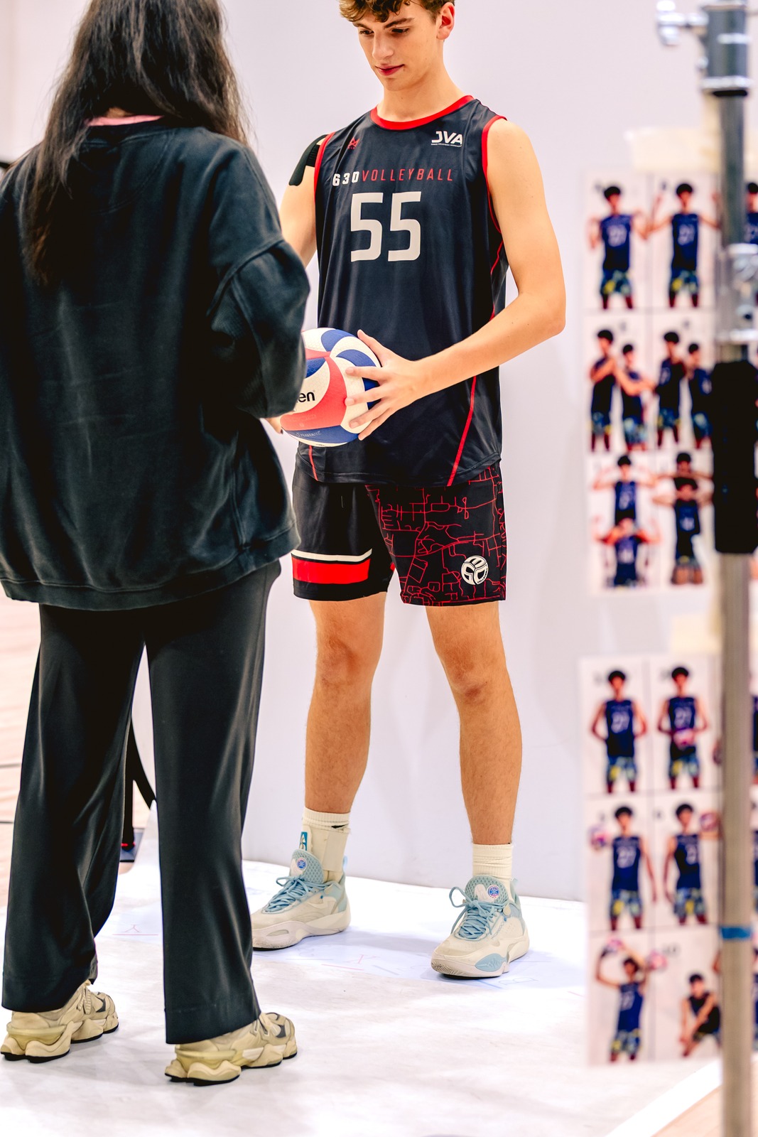 A player in a dark jersey holds a volleyball beside a person in black at the photo setup.