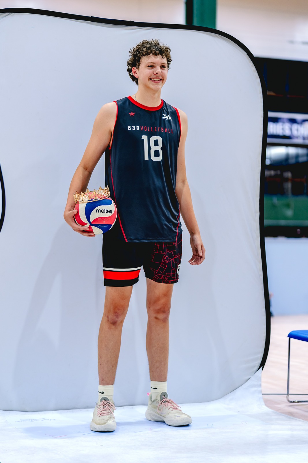 A smiling player in a dark blue jersey and black shorts stands holding a volleyball with a crown on top.