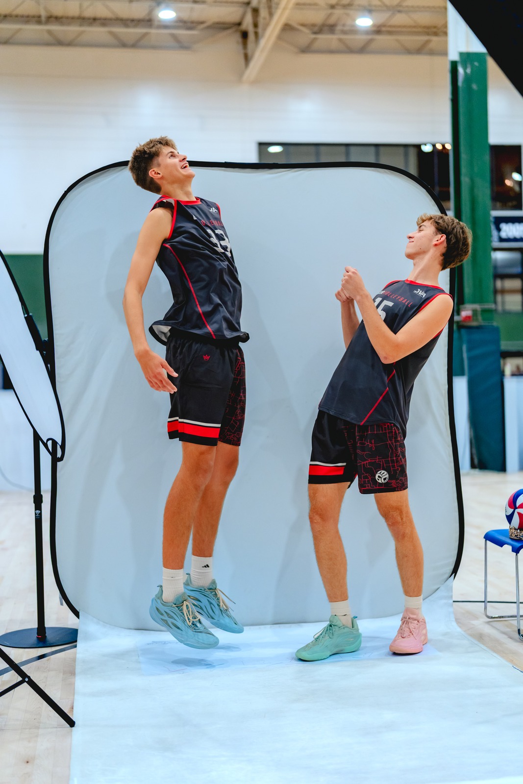 One player in black and red jumps beside another with hands raised at a photo backdrop.