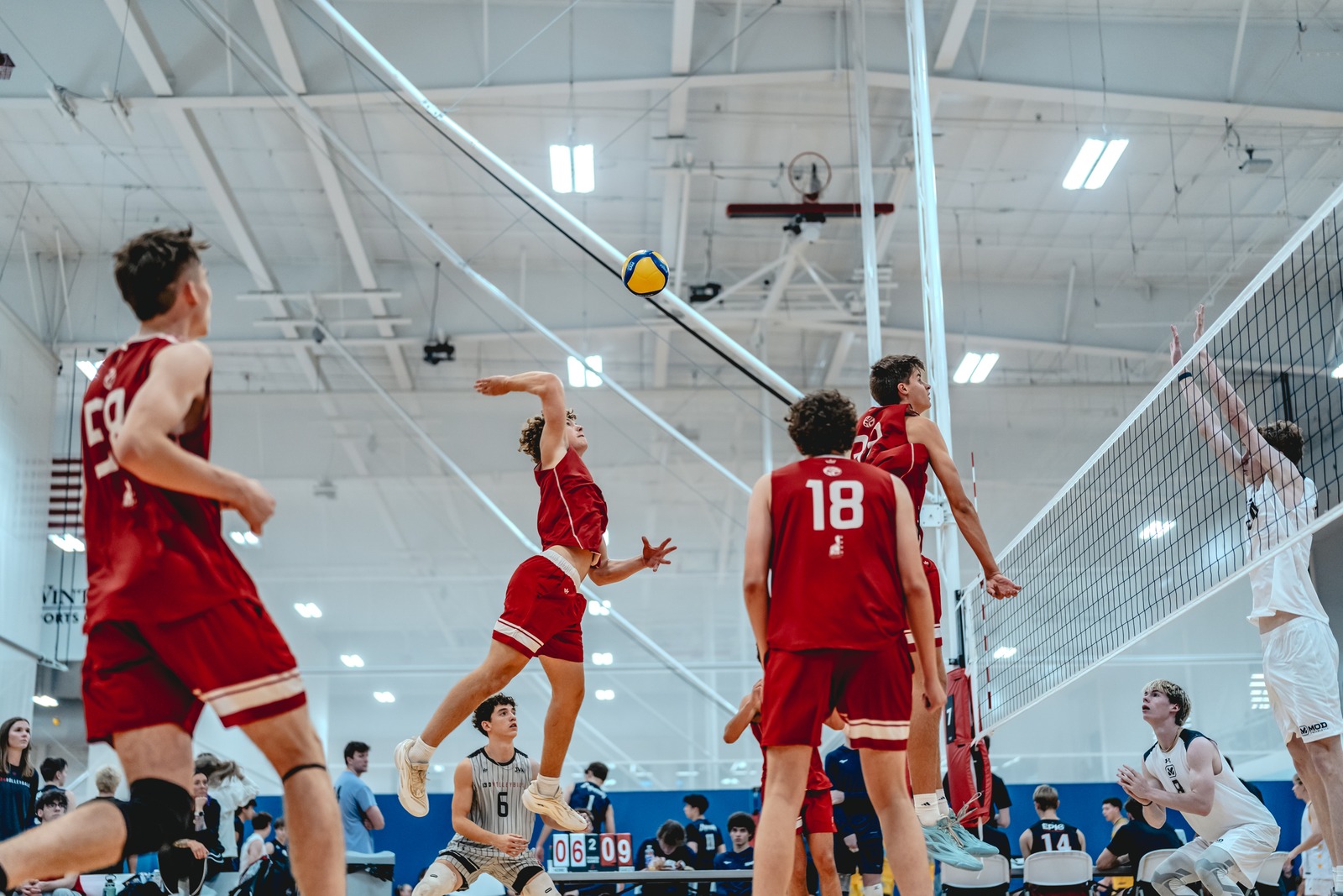 A player in red jumps with arms extended, while another player in red stands nearby and a player in white is at the net.