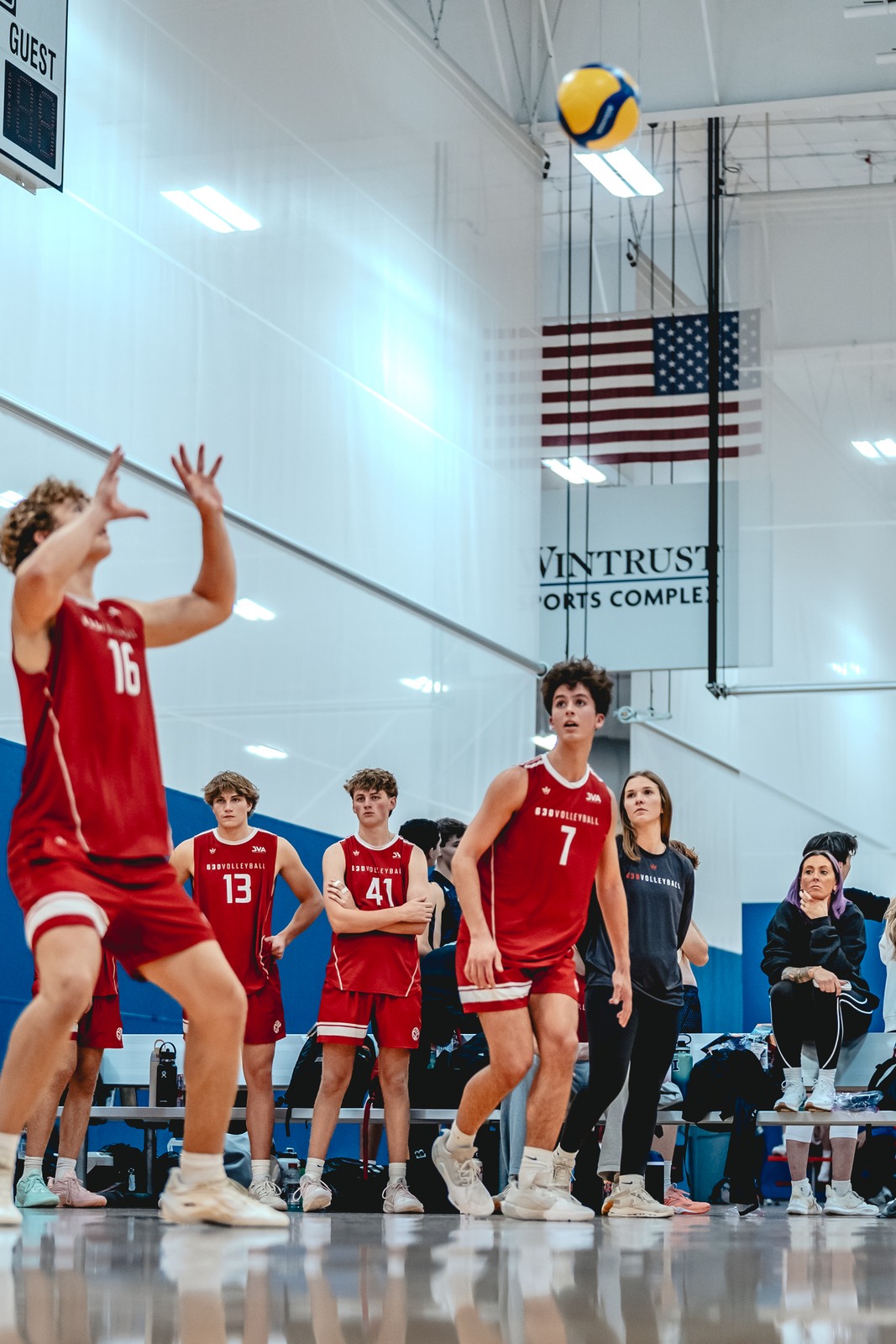 A player in red reaches up with hands open as a ball flies overhead.