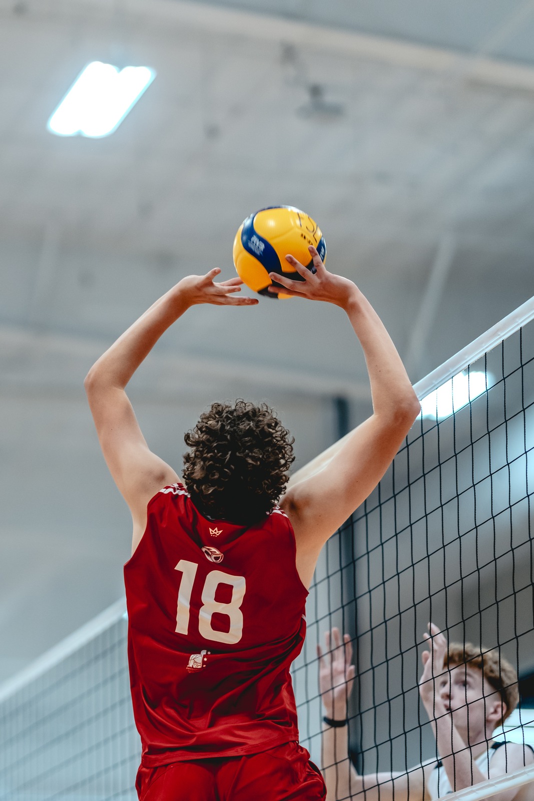A player in red reaches overhead with a ball, while a player in white extends hands forward.