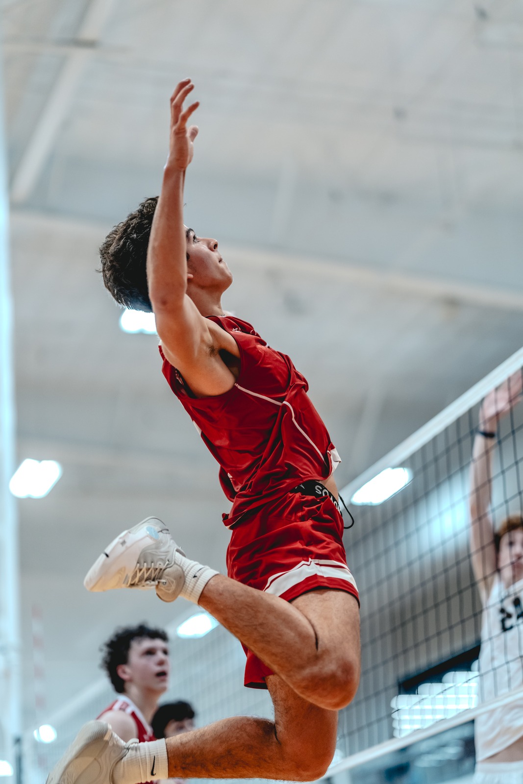 A player in a red top and shorts jumps with one arm extended upwards. Another player in a white top is visible behind the net.