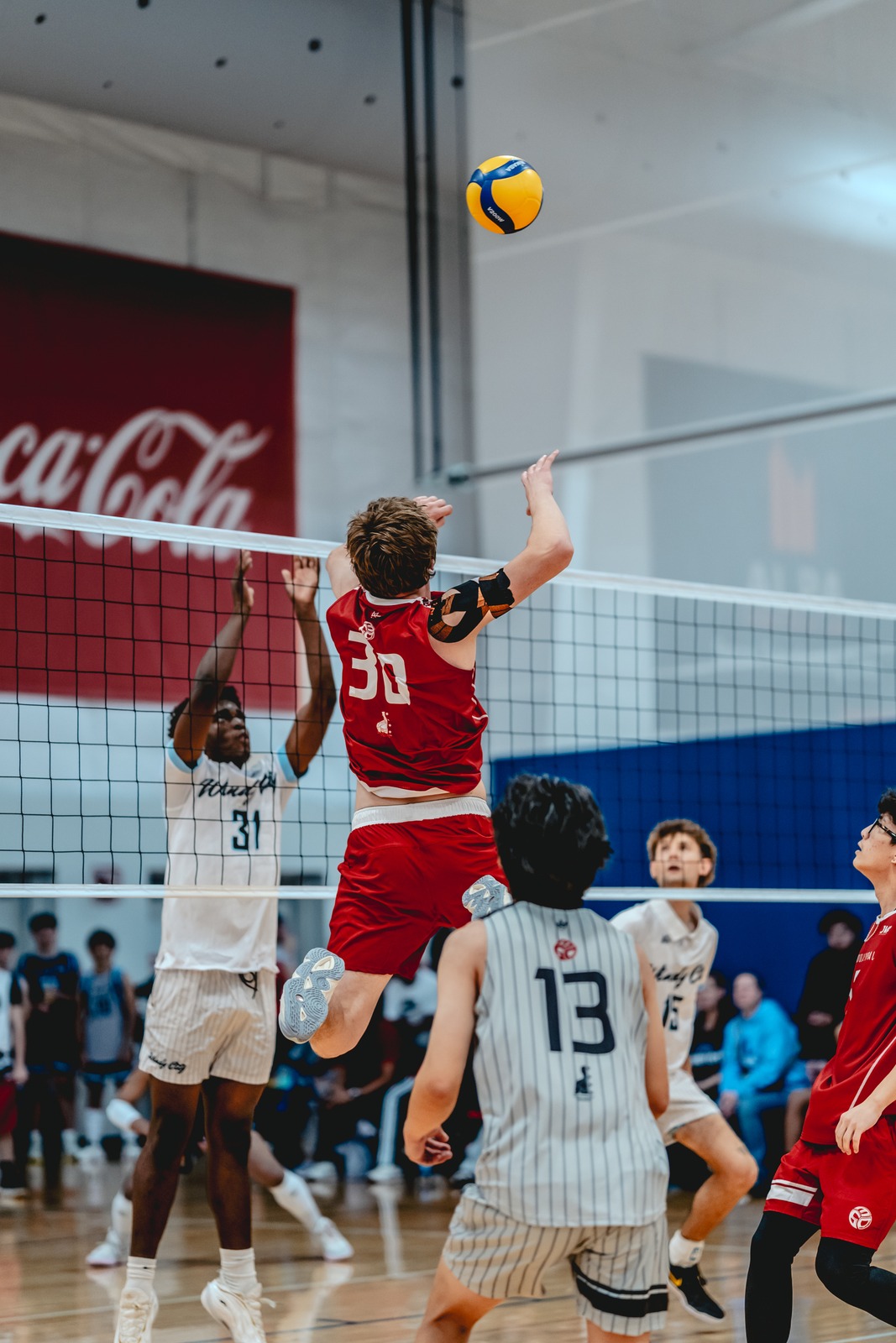 A player in red jumps with arms extended toward the ball. A player in white reaches up. A player in gray watches.