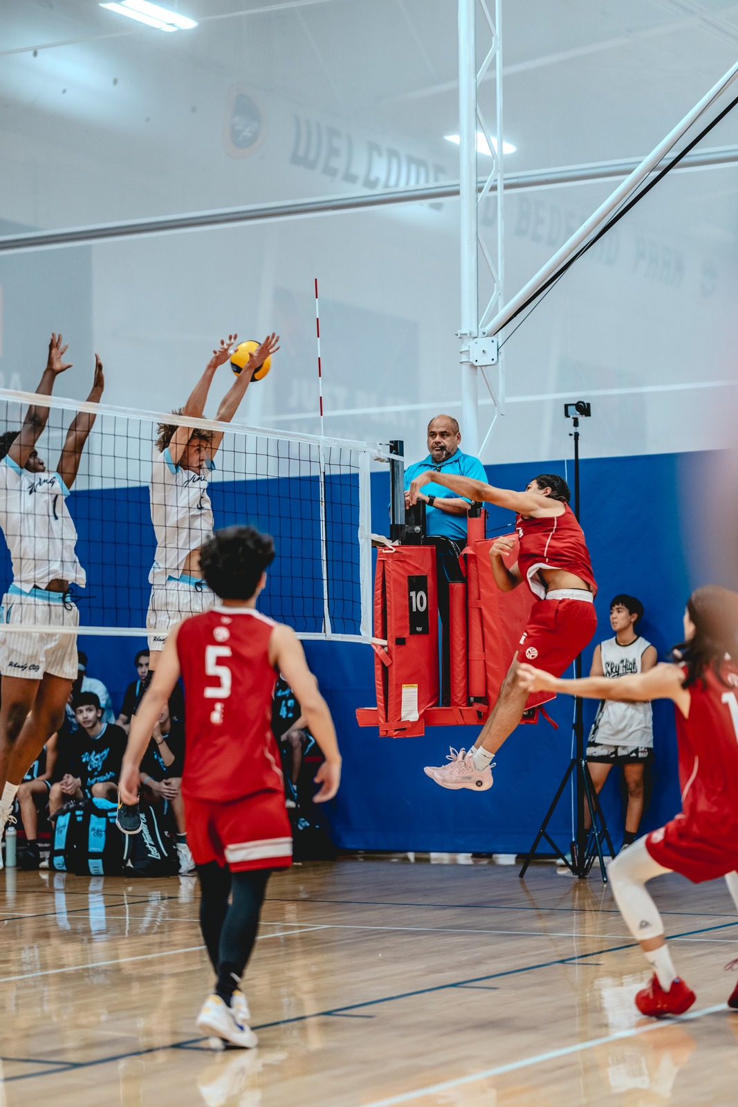 A player in red jumps with arms extended, reaching for the ball. Another player in white reaches up.