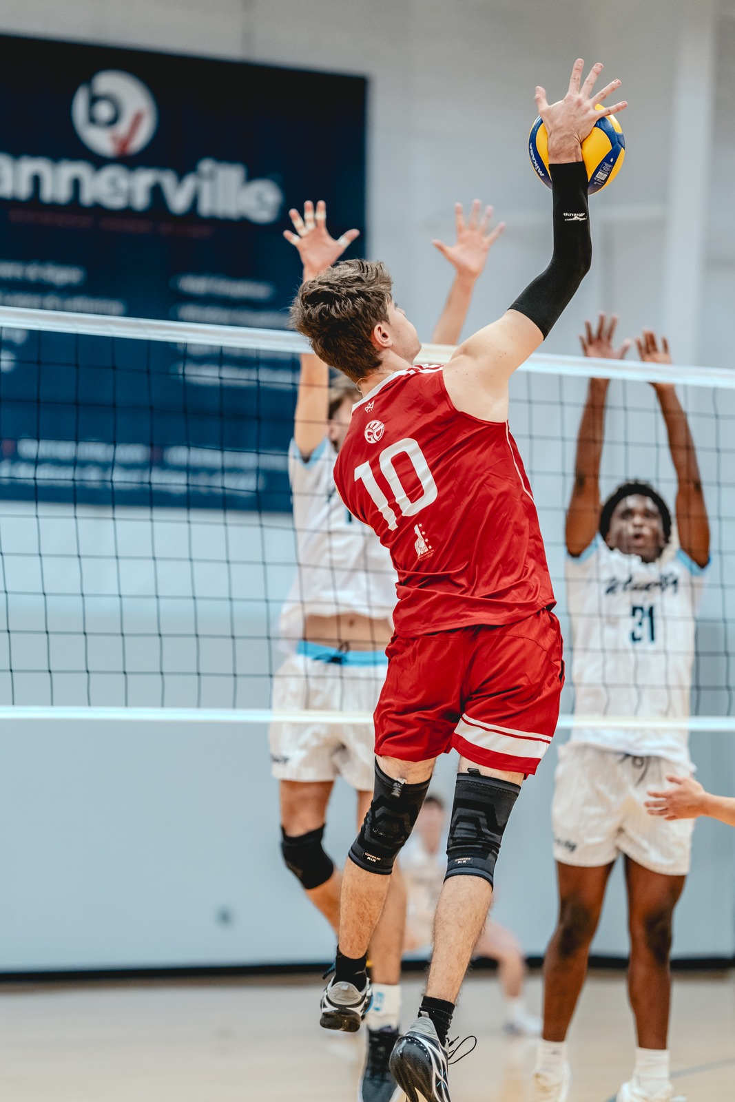 A player in a red top reaches overhead for the ball while players in white tops jump with hands raised.