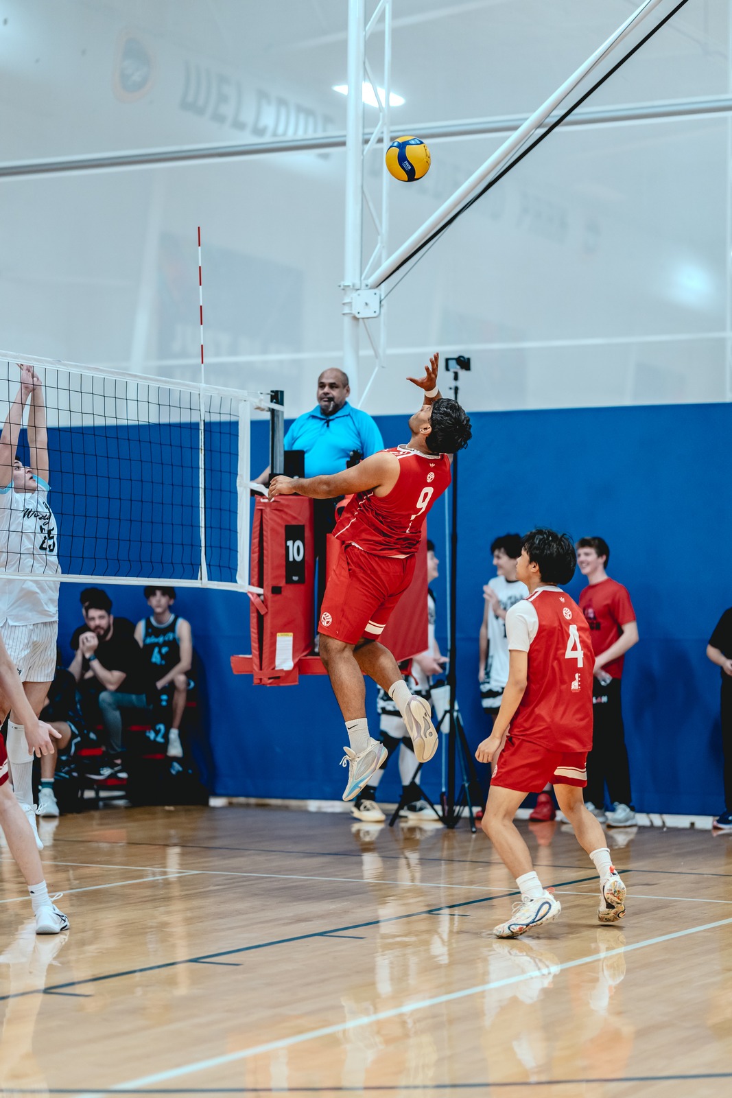 A player in red reaches overhead for the ball while another player in white reaches up.