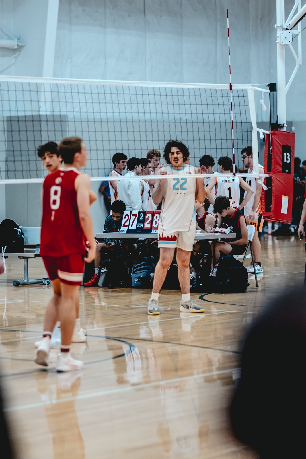A player in a red jersey stands on a court, while a player in a white jersey with blue trim stands facing forward.